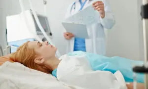 Pacjentka leży w łóżku szpitalnym, podłączona do respiratora. Lekarz analizuje wyniki badań, monitorując zachowanie po wybudzeniu ze śpiączki farmakologicznej.