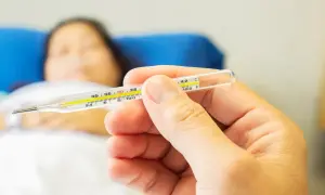 Dłoń trzyma termometr, pokazując temperaturę. W tle leży chora osoba, która potrzebuje pomocy, jak zbić gorączkę.