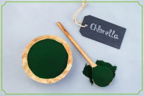 Zielony proszek w drewnianej miseczce i na łyżce. Tabliczka z napisem "Chlorella" wyjaśnia, co to jest chlorella.