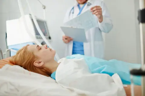 Pacjentka leży w łóżku szpitalnym, podłączona do respiratora. Lekarz analizuje wyniki badań, monitorując zachowanie po wybudzeniu ze śpiączki farmakologicznej.