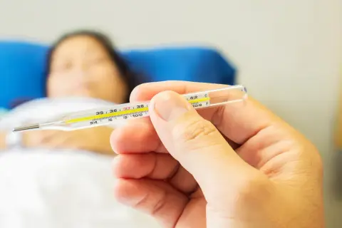 Dłoń trzyma termometr, pokazując temperaturę. W tle leży chora osoba, która potrzebuje pomocy, jak zbić gorączkę.