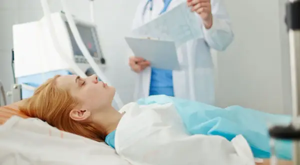 Pacjentka leży w łóżku szpitalnym, podłączona do respiratora. Lekarz analizuje wyniki badań, monitorując zachowanie po wybudzeniu ze śpiączki farmakologicznej.