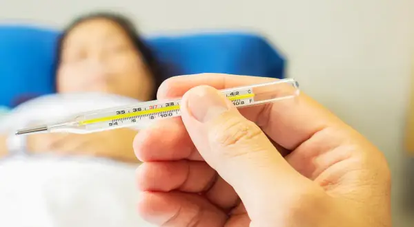 Dłoń trzyma termometr, pokazując temperaturę. W tle leży chora osoba, która potrzebuje pomocy, jak zbić gorączkę.
