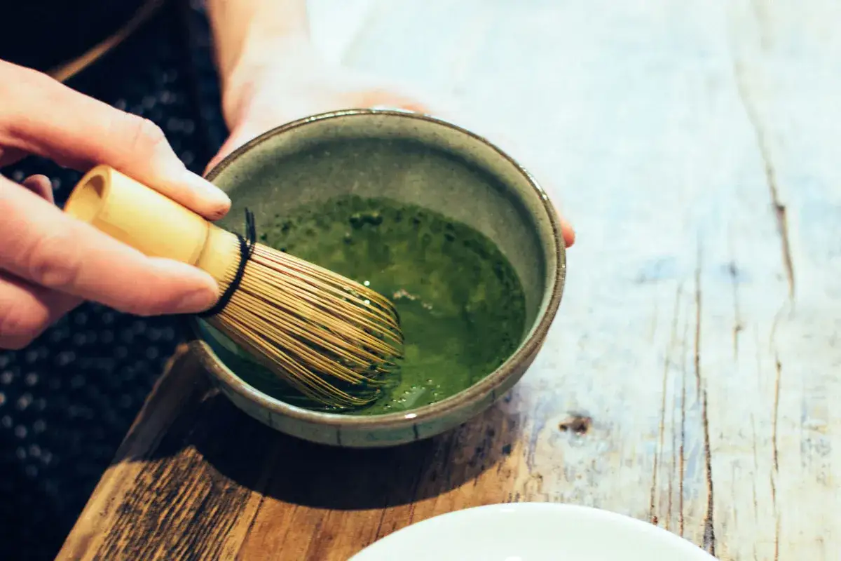 Ręce ubijają bambusową miotełką zielone matcha do picia w ceramicznej miseczce.